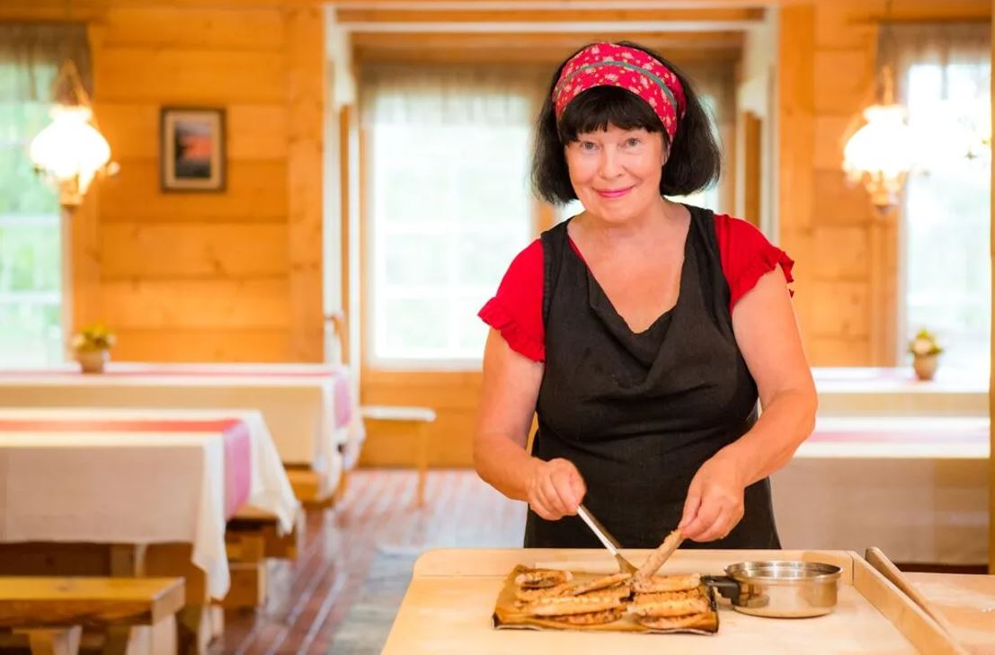 Hymyilevä nainen voitelee vastapaistettuja karjalanpiirakoita tuvassa. 